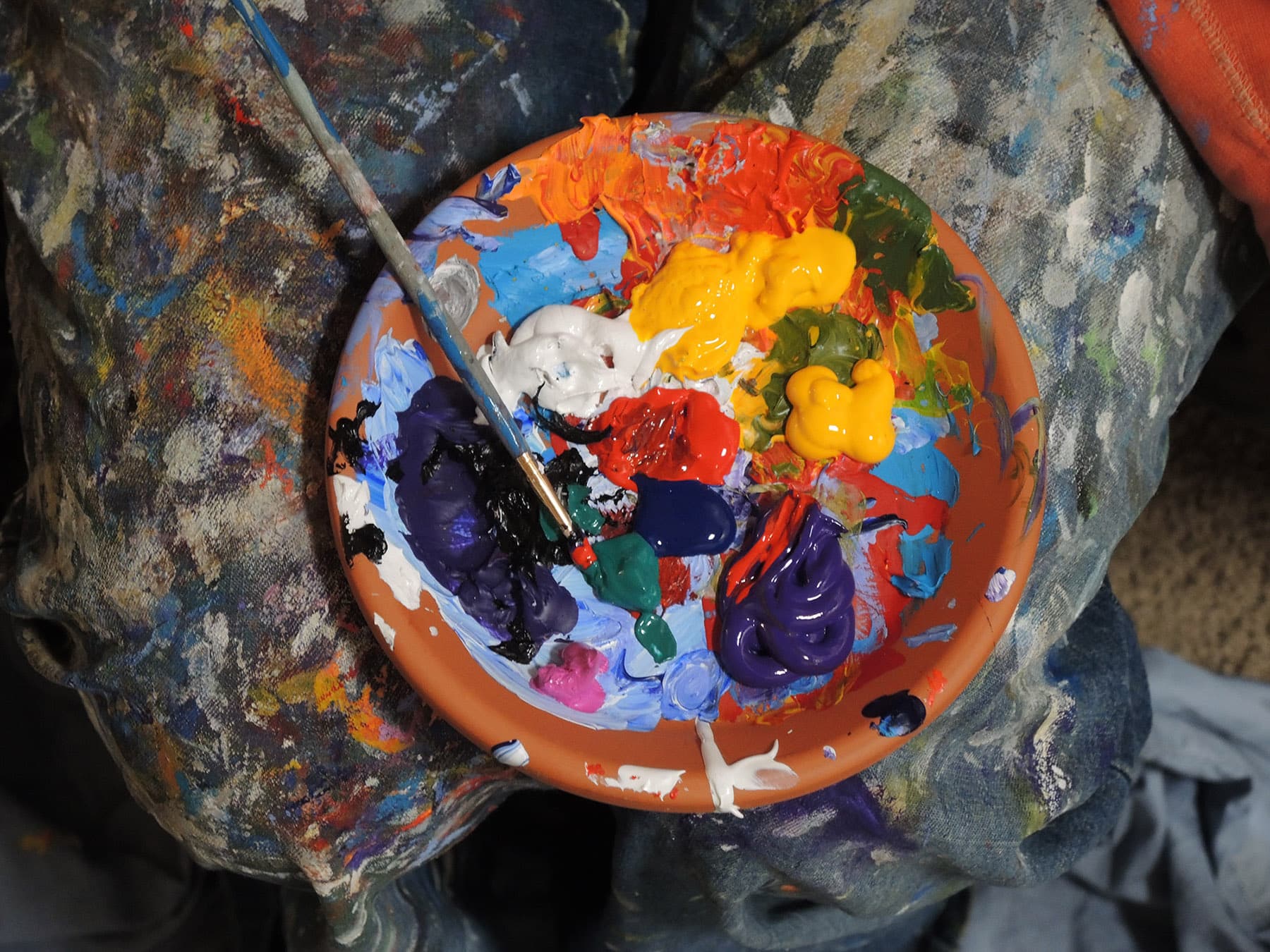 A shallow bowl on a very used paint stained surface. There are various colors of paint in the bowl with a paint brush left laying in it.